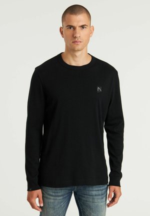 Camiseta de manga larga negra de algodón con cuello redondo, que presenta un pequeño parche de logo en el pecho y puños acanalados.