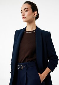 Marineblazer met een gladde textuur, afgelegd over een bruine gebreide top met een zilveren accent bij de halslijn, gecombineerd met een op maat gemaakte broek.