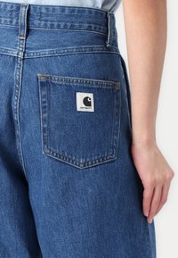 Siniset denimmekot ovat korkeavyötäröiset, niissä on takatasku, jossa on valkoisella ommeltu logopatchi, ja suoralahkeinen malli, jossa on vähäistä kulutusta.