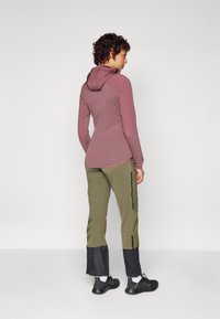Haut à manches longues avec capuche en tissu maroon clair, associé à un pantalon vert olive avec des fermetures éclair et des accents noirs ; design athlétique épuré.