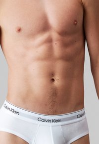 Boxers en coton blanc avec une ceinture portant un logo, offrant un ajustement serré et une texture douce. L'abdomen bien dessiné du modèle est clairement visible.