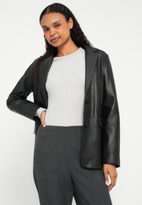 Μαύρο σακάκι από ψεύτικο δέρμα με γιακάδες με εγκοπές, δύο κουμπιά και πλαϊνές τσέπες, φορεμένο πάνω από μια ανοιχτογκρι ριγωτή πουλόβερ και σκούρες γκρι παντελόνες.