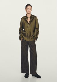 Camicia verde in satin con colletto, abbinata a pantaloni marroni larghi. La camicia ha maniche lunghe e una leggera lucentezza. Calzature nere.