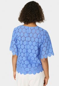 Top in pizzo blu con motivi floreali, maniche corte a scallop e zip sulla schiena. Tessuto leggero con una finitura testurizzata.