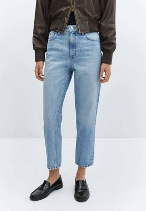 Person iført lyseblå højtaljede jeans kombineret med sorte loafers og en kort brun læderjakke mod en ensfarvet hvid baggrund.