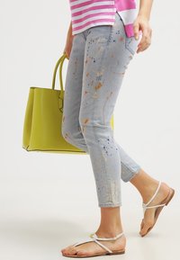 Persona que lleva unos vaqueros de mezclilla de color azul claro salpicados de pintura, sandalias blancas, una camiseta a rayas rosas, y sostiene un gran bolso de color amarillo-verde brillante mientras camina.