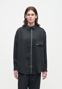 Μαύρο σατέν πουκάμισο με κουμπιά και λευκή διακόσμηση περιμετρικά, μία τσέπη στο στήθος και μικρό λογότυπο στην τσέπη. Μακριά μανίκια και άνετη εφαρμογή.