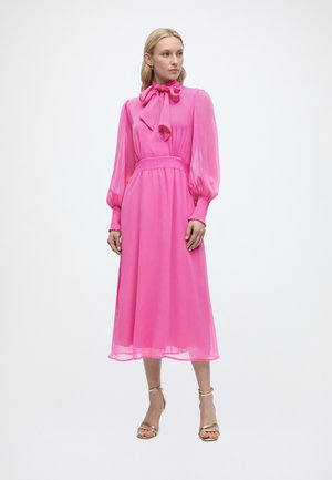 Femeie în picioare, purtând o rochie midi roz strălucitor cu mâneci lungi, o fundă mare la gât și pantofi cu toc aurii cu bretea la gleznă.