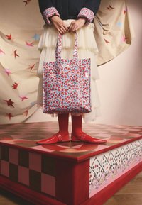 Gėlių raštais puoštas pirkinių krepšys su raudonomis ir mėlynomis gėlėmis šviesiai mėlyname fone; modelis dėvi raudonus batus ir kremine sluoksniuota sijoną. Raudonos detalės.