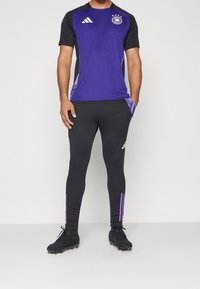 Maglietta sportiva viola con accenti neri, maniche corte e tre stelle. Leggings neri con strisce viola ai lati. Scarpe da calcio nere.