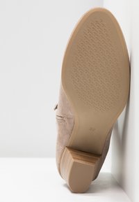Semelle de chaussure en daim beige avec un talon bloc, taille 37, reposant verticalement contre un mur et un sol blancs.