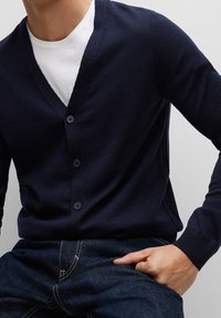 Námořnicky modrý pletený kardigan s výstřihem do V a pěti knoflíky vpředu. Nošený přes bílé tričko, v kombinaci s tmavě modrými džínami.