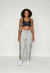 Svart sportsbh med tre hvite striper, grå joggebukser med svarte sidestriper, og hvite joggesko. Modellen står foran en nøytral bakgrunn.