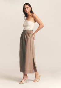 Vaalean beige ribbitekoinen hihaton toppi, yhdistettynä korkeavyötäröiseen taupe-maksihameeseen, jossa on röyhelövyötärö ja sivuhalkio. Beige korkokenkiä.