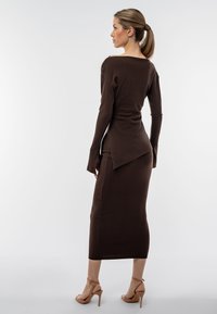 Vestito a coste marrone con maniche lunghe e un orlo asimmetrico, caratterizzato da una silhouette aderente e spacchi laterali, abbinato a tacchi con cinturini.