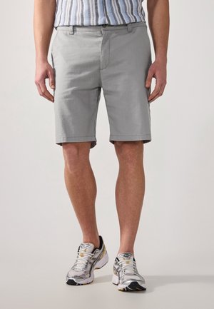 Homme portant un short gris clair à hauteur des genoux, une chemise rayée rentrée et des chaussures de course argentées avec des touches jaunes.