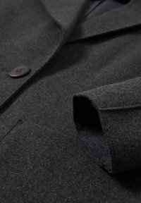 Manche de manteau en laine gris foncé avec devant boutonné, couture visible et détail de poche sur tissu texturé.