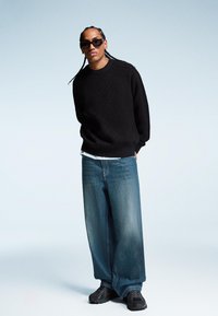 Črn pulover z teksturo, ohlapne svetlo modre denim hlače in temni športni copati. Model ima spletene lase in nosi temna sončna očala.