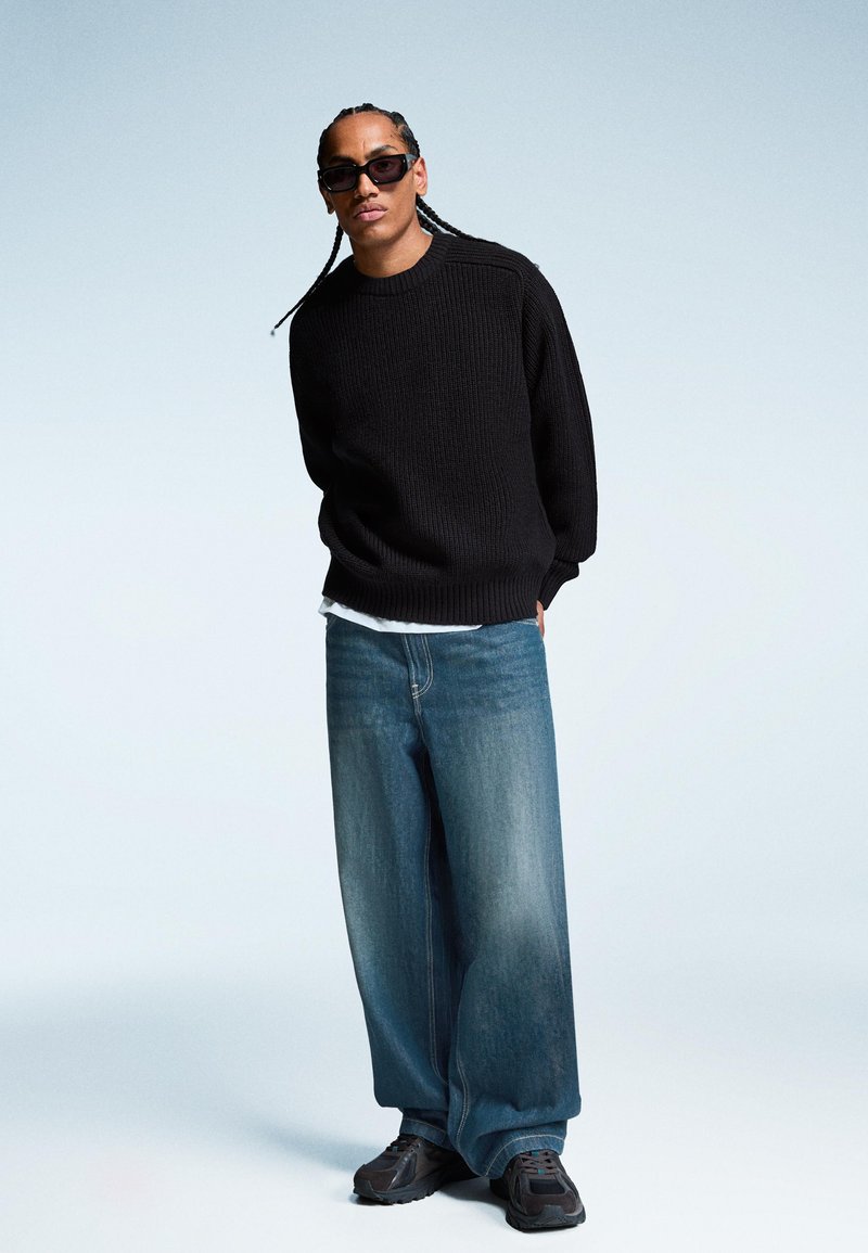 Črn pulover z teksturo, ohlapne svetlo modre denim hlače in temni športni copati. Model ima spletene lase in nosi temna sončna očala.