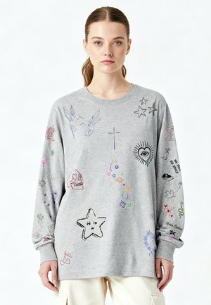 Giovane donna con capelli castani raccolti in una coda di cavallo che indossa una felpa grigia decorata con piccoli disegni e simboli multicolori in stile schizzo.