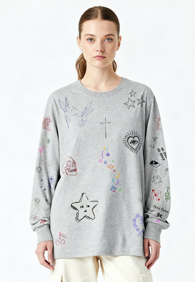Jeune femme aux cheveux bruns attachés en queue de cheval, portant un sweat-shirt gris orné de petits motifs et symboles multicolores de style griffonné.