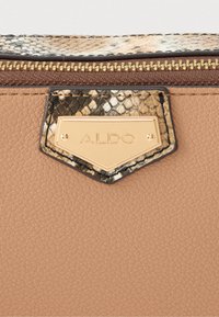 Světle hnědá kožená taška s texturou, zlatou kovovou destičkou s logem a hadím motivem jako akcentem. Na horním okraji je viditelný zip.