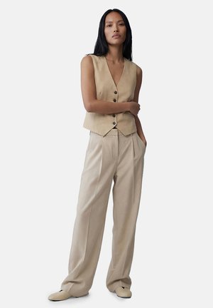 Kvinde stående med armene over kors iført beige knappet vest, beige bukser med vide ben og matchende sko mod en ensfarvet lys baggrund.