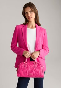 Lyserød quiltet håndtaske med guld hardware, rund form, top-hænder og mærkets logo. Båret med en lys pink blazer og hvid top.
