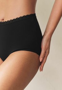 Augsta vidukļa melnas kokvilnas biksītes ar mežģīņu apdari jostasvietā un gludu tekstūru, redzamas no sāniem ar roku uz gurna.