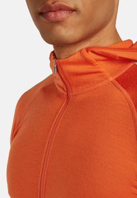 Primer plano de una persona que lleva una chaqueta naranja con cremallera y un alto cuello, mostrando la parte inferior de la cara y el cuello.