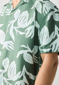 Kortermet grønn skjorte med hvitt blomstermønster, med krage og lyse knapper. Stoffet har en jevn, lett strukturert overflate.