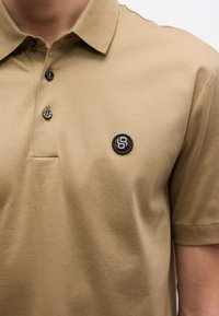 Krátke polo tričko s krátkym rukávom z hladkej látky, s trojprvkovým zapínaním na gombíky a okrúhlym čiernym logom na ľavej strane hrudníka.