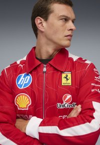 Giovane in tuta da corsa rossa Ferrari con le braccia incrociate, che guarda di lato, mostrando i loghi di HP, Shell, UniCredit e Ferrari.