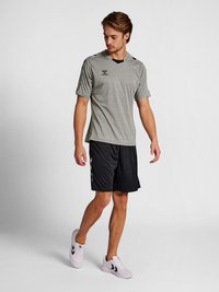Jeune homme portant un t-shirt de sport gris, un short noir et des baskets blanches, debout et regardant vers le bas à sa gauche.