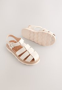 Sandale blanche avec une semelle intérieure en liège, présentant un design à brides croisées, une boucle de cheville réglable et une semelle extérieure en caoutchouc texturée pour une meilleure adhérence.