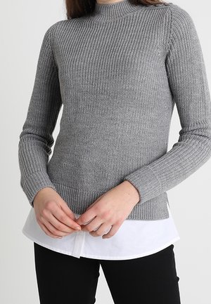 Kobieta ubrana w szary prążkowany sweter na biały shirt i czarne spodnie, ręce splecione przed sobą.