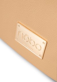 Beige strukturiertes Kunstlederhandtasche mit einem goldfarbenen Markenplakette mit eingraviertem Text und dekorativen Hardwareakzenten.