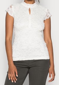 Haut en dentelle blanche avec un col montant, des manches courtes et un détail en forme de clé. motif floral texturé avec des bords festonnés aux manches et à l'ourlet.