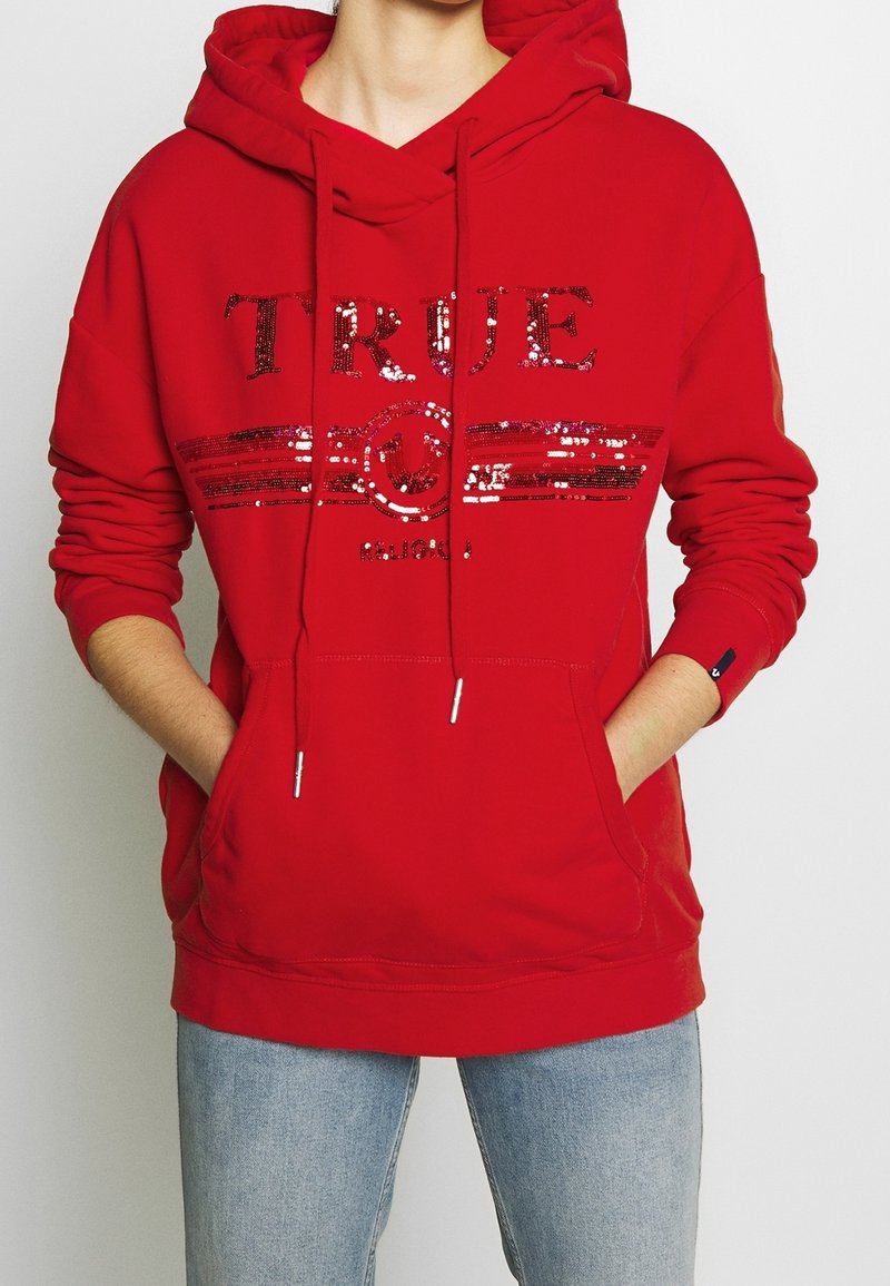 Personne portant un sweat à capuche rouge avec le texte "TRUE" en sequins et un design, mains dans les poches avant, associée à un jean bleu clair.