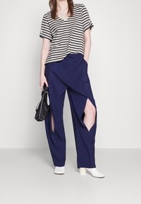 Camisa de manga corta a rayas en beige y negro; pantalones azul marino con detalles recortados; zapatos de tacón bloque blancos; bolso negro pequeño con rayas.