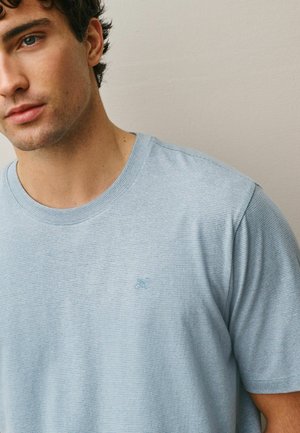 Junger Mann trägt ein hellblaues, fein gestreiftes T-Shirt mit einem kleinen gestickten Logo auf der Brust vor einfachem beigen Hintergrund.