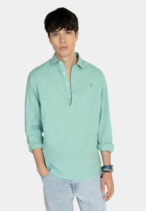 Junger Mann mit dunklen Haaren, der ein mintgrünes, langärmeliges Hemd mit Knöpfen und hellblaue Jeans trägt, mit Armbändern am Handgelenk.