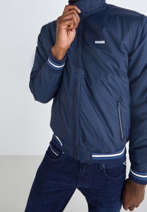 Un homme ferme une veste aviateur bleu marine à rayures aux poignets et à la taille, portant un jean foncé, sur un fond clair uni.