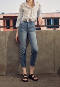 Jeans cortos de color azul claro con una cintura alta, con el borde deshilachado, combinados con sandalias negras. Camisa blanca de botones anudada a la cintura.