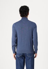 Texturerad blå stickad tröja med hög krage, långa ärmar och ribbade manschetter, i kombination med denimjeans. Bakifrån visas en enkel design.