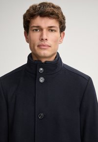 Marineblauer Wollmantel mit hohem Kragen, ausgestattet mit drei vorderen Knöpfen und einer glatten Textur. Einfaches, strukturiertes Design mit verdecktem Reißverschluss.
