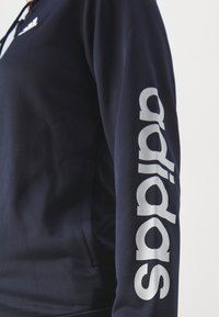 Námořnická mikina s hladkou texturou a dlouhými rukávy. Je zdobena bílým logem "adidas", které je výrazně umístěno po celé délce rukávu.