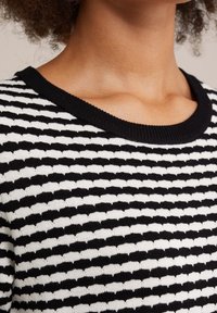 Pull en tricot rayé noir et blanc avec un col côtelé et un tissu texturé, présentant des motifs alternés créant un effet de vague.