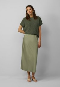 Haut en tricot vert à manches courtes, associé à une longue jupe maxi légère de couleur vert clair. Le modèle porte des sandales à lanières beiges. Design simple et texturé.