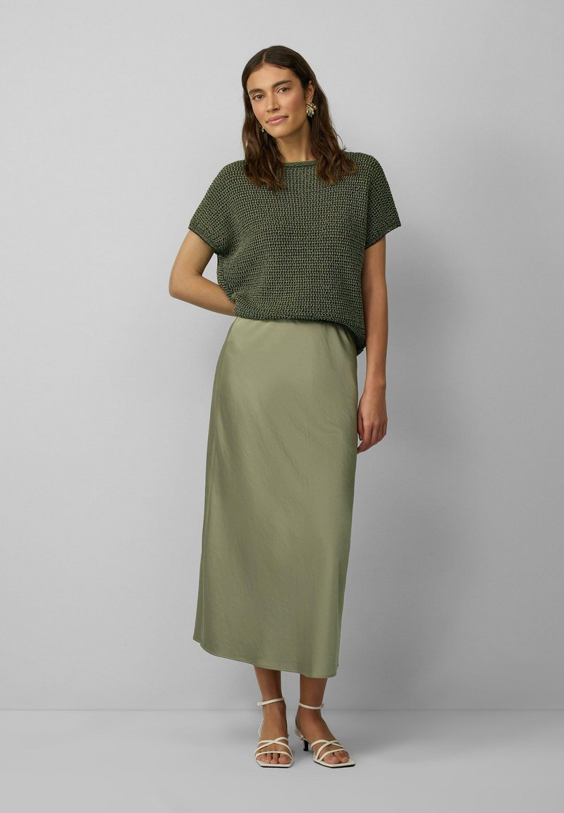 Haut en tricot vert à manches courtes, associé à une longue jupe maxi légère de couleur vert clair. Le modèle porte des sandales à lanières beiges. Design simple et texturé.